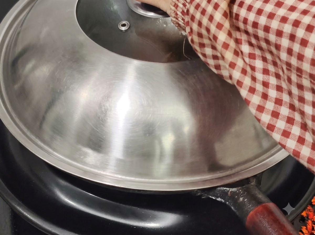 A metal wok lid covering the wok on a stovetop, being held by a hand in a red and white checkered sleeve.