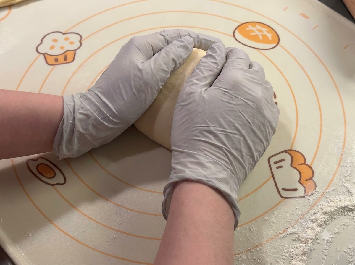 Gloved hands shaping and rounding dough portions on a baking mat.
