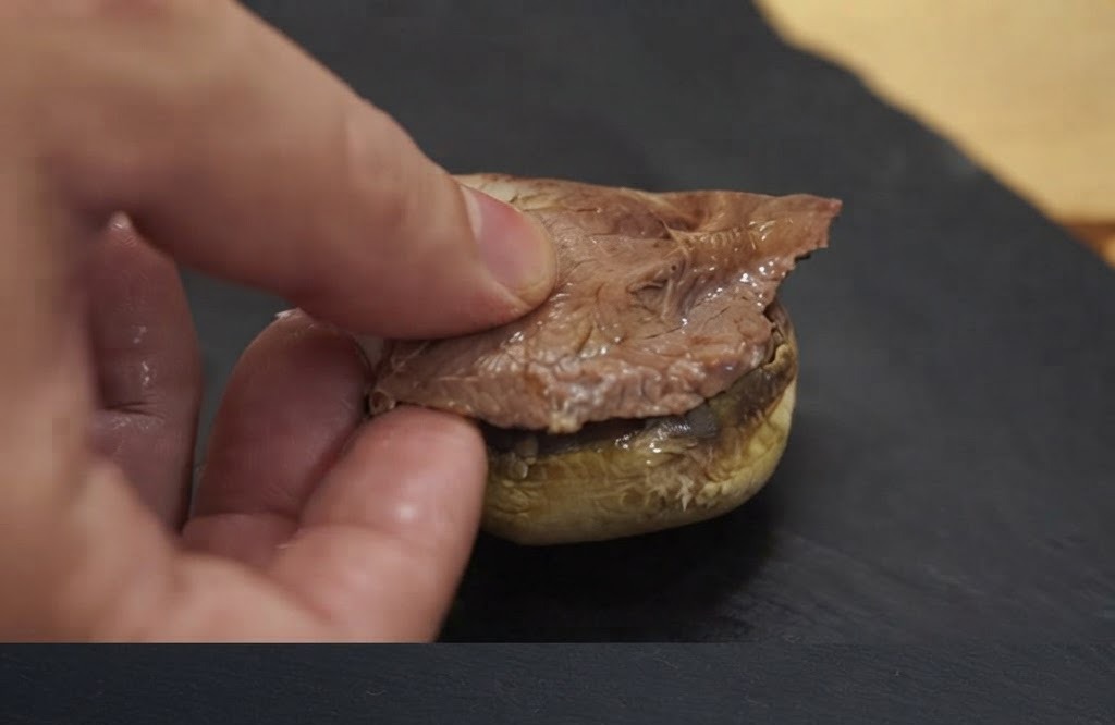 A hand placing a piece of cooked meat onto a baked mushroom cap base.
