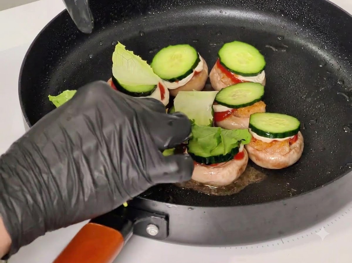 A gloved hand placing toothpicks through stacked ingredients in mushroom caps inside a black frying pan.