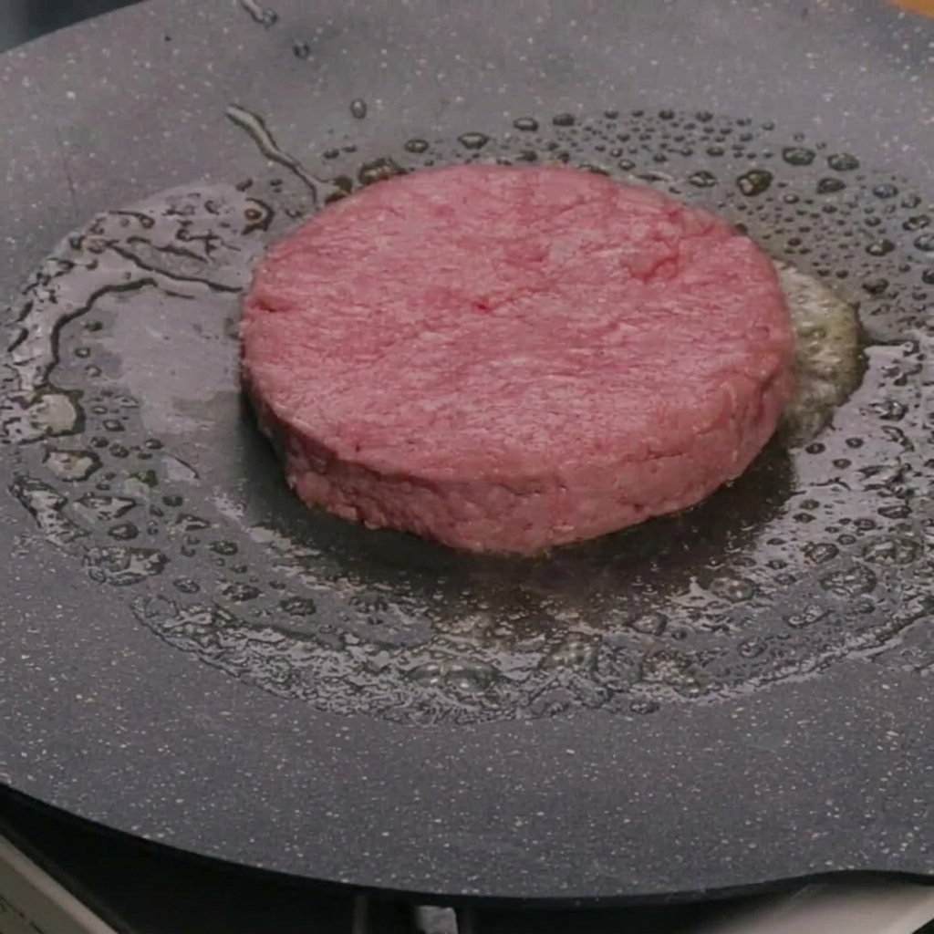 A raw beef patty cooking in butter inside a hot frying pan.