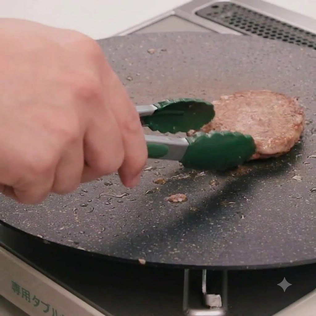 A hand using green kitchen tongs to flip a beef patty cooking in a flat skillet.