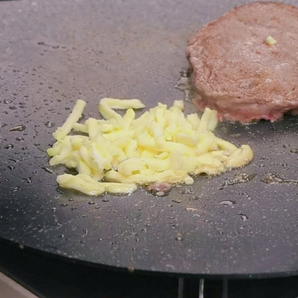 Shredded mozzarella cheese melting on top of a pan-fried beef patty in a hot skillet.