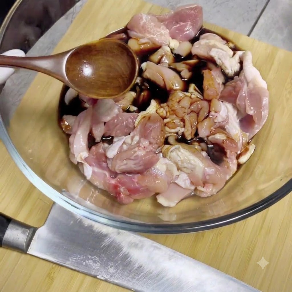 Soy sauce being poured from a wooden spoon onto raw chicken pieces inside a glass mixing bowl.