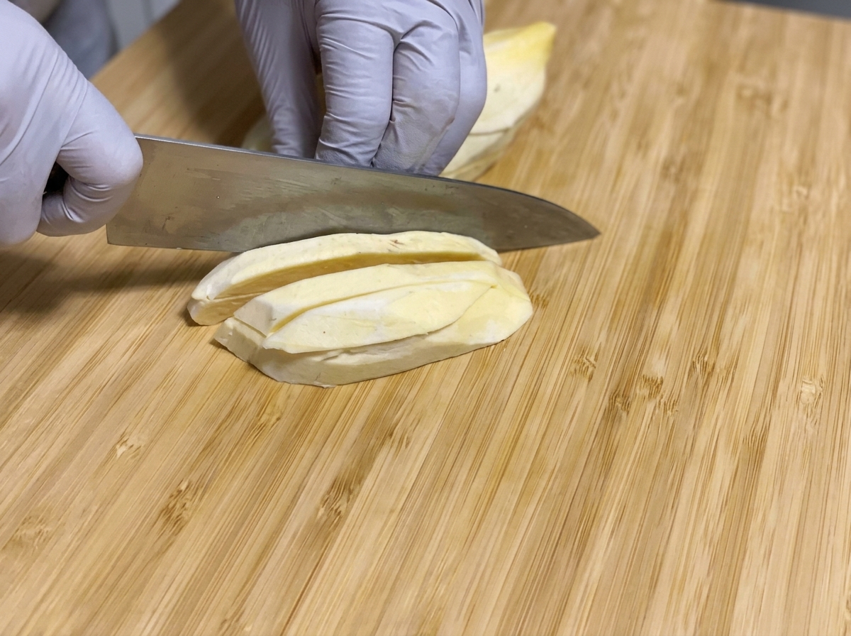 Peeled raw sweet potatoes being sliced into long sticks with a knife on a wooden cutting board.