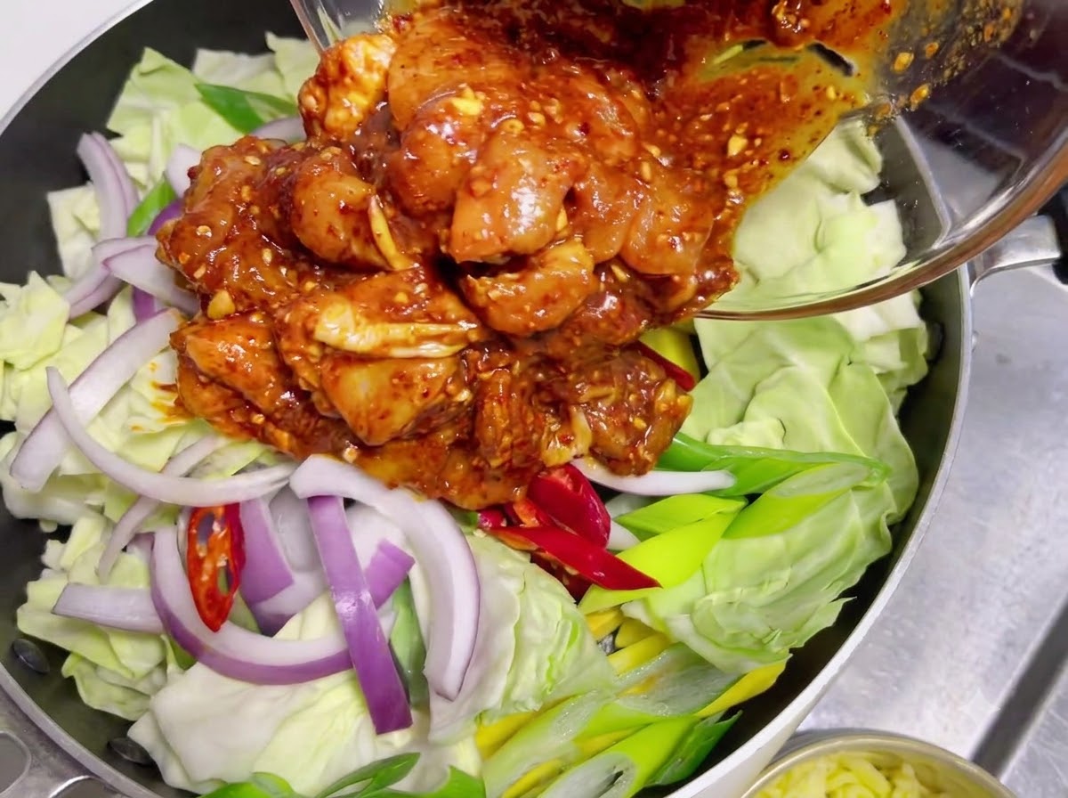 Marinated raw chicken coated in spicy red sauce being poured from a glass bowl onto a layer of raw cabbage and onions.