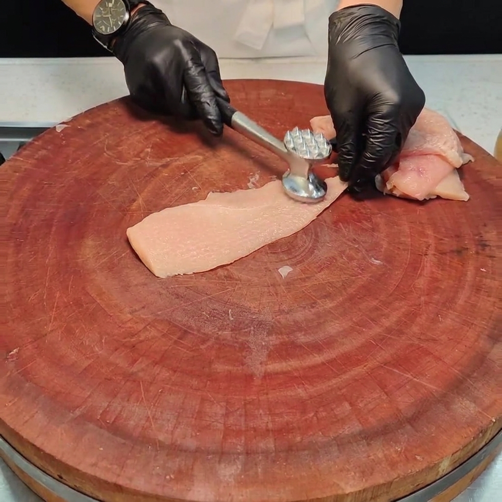 A piece of chicken breast being pounded flat with a metal meat mallet on a wooden board.