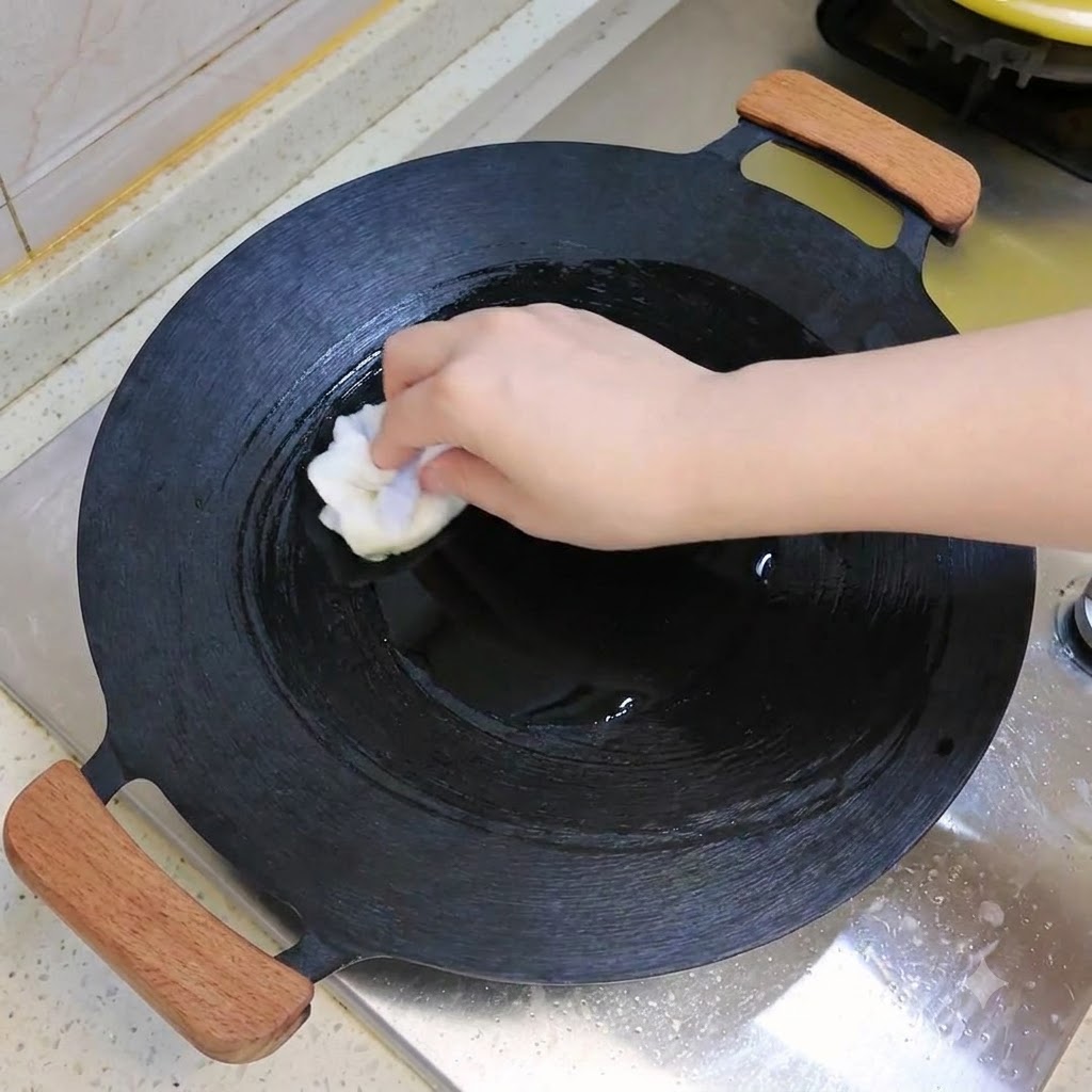 A dark wok being wiped or oiled with a white cloth on a stovetop.