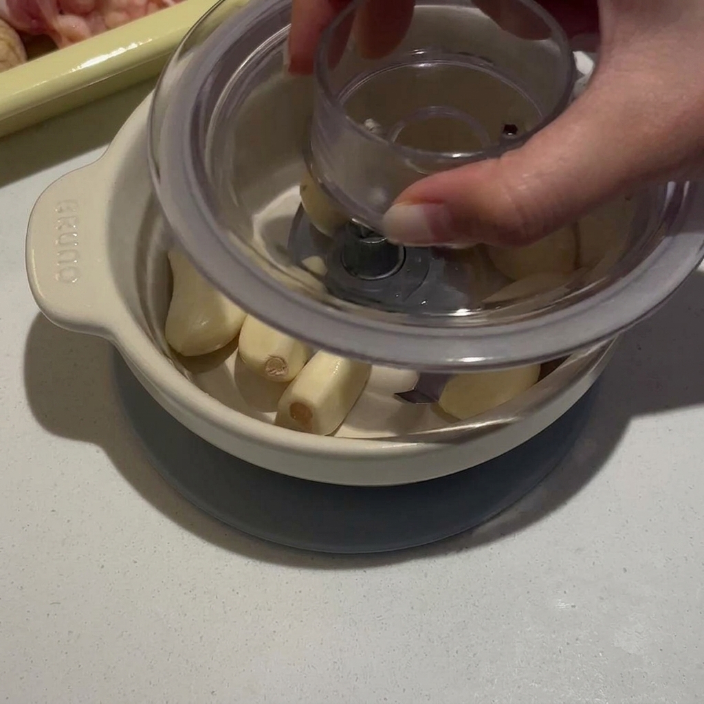 A person pressing down on the lid of a small manual food processor filled with peeled garlic cloves.