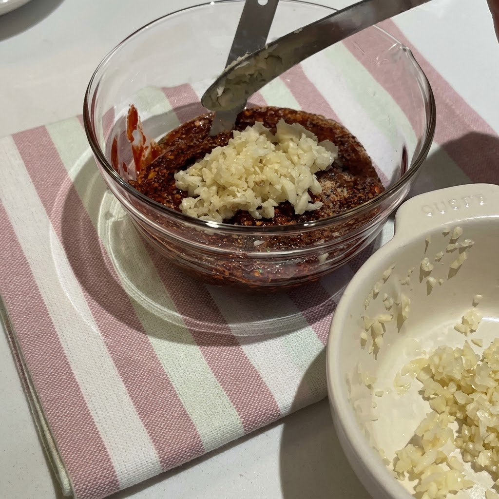 Korean chili paste and various seasonings being mixed in a glass bowl to create a thick red sauce.