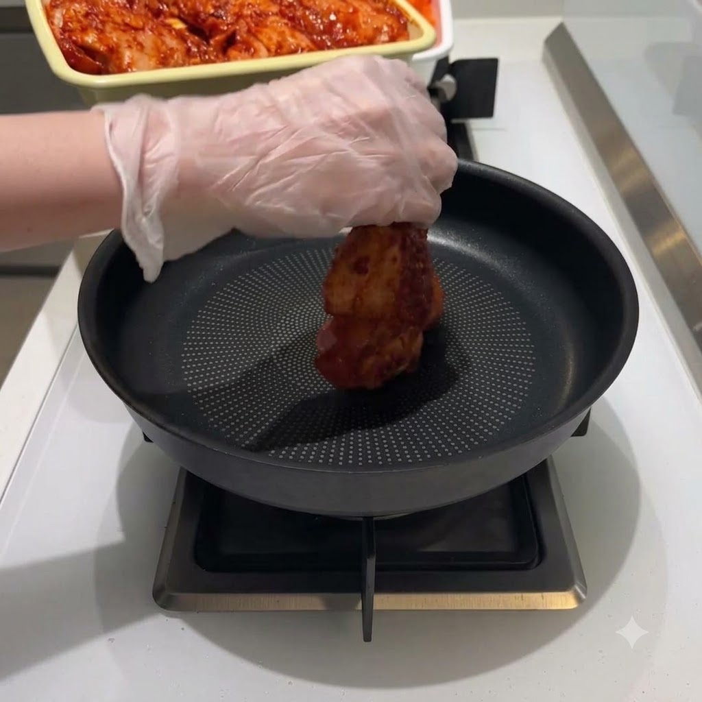 A gloved hand placing a piece of red marinated chicken into the middle of a black frying pan.