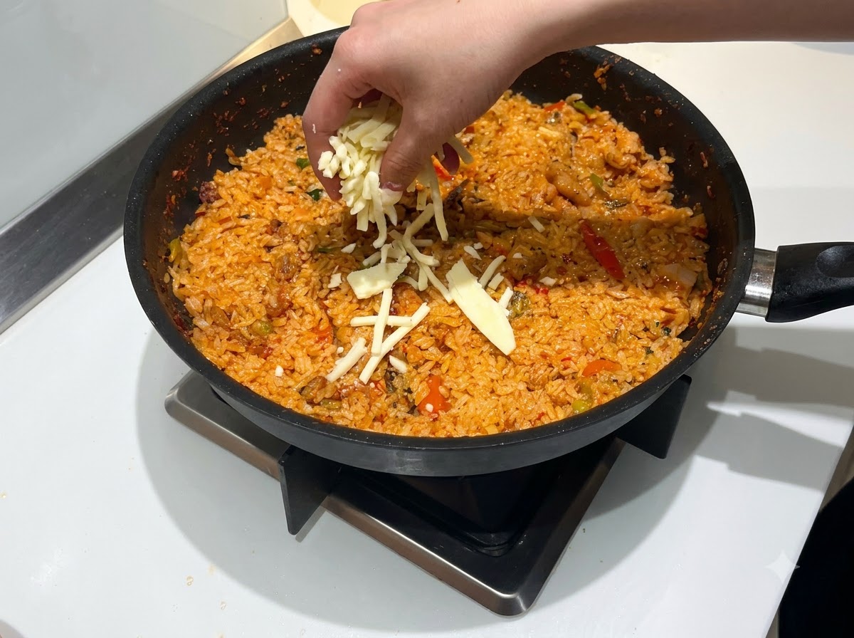 A hand sprinkling a generous amount of shredded mozzarella cheese over a pan of red, spicy fried rice.