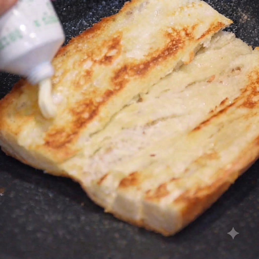 Mayonnaise being squeezed from a tube onto a golden toasted baguette half.
