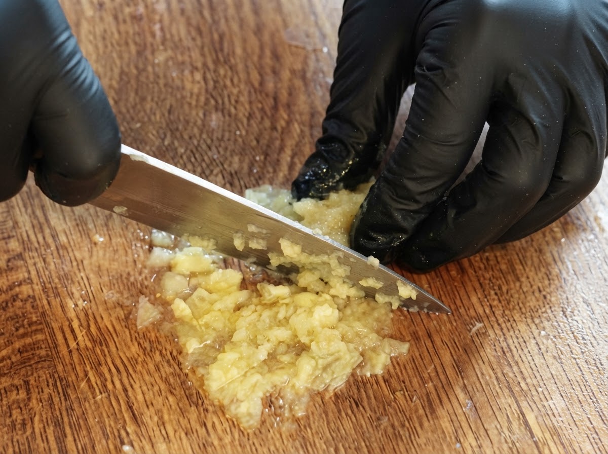 A gloved pair of hands using a knife to chop and puree leftover lemon flesh on a wooden cutting board.