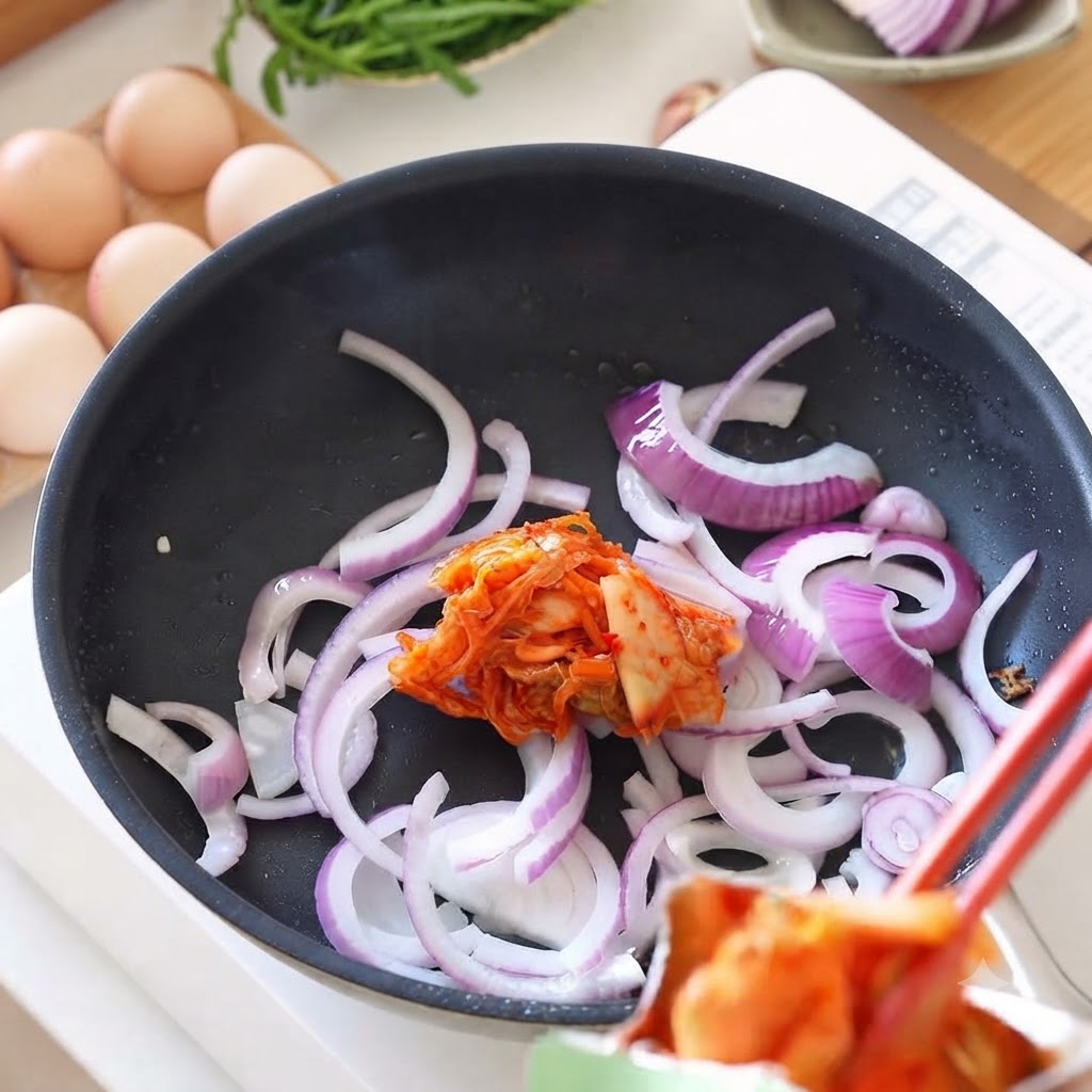 Fresh kimchi being added from chopsticks into a pan containing sautéed onions.