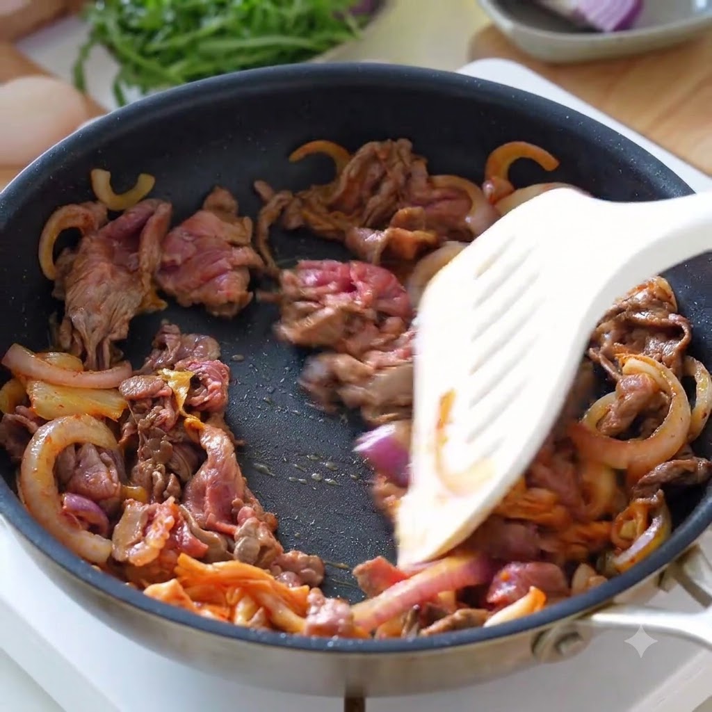 Sliced beef, onions, and kimchi being tossed together in a frying pan with a spatula.