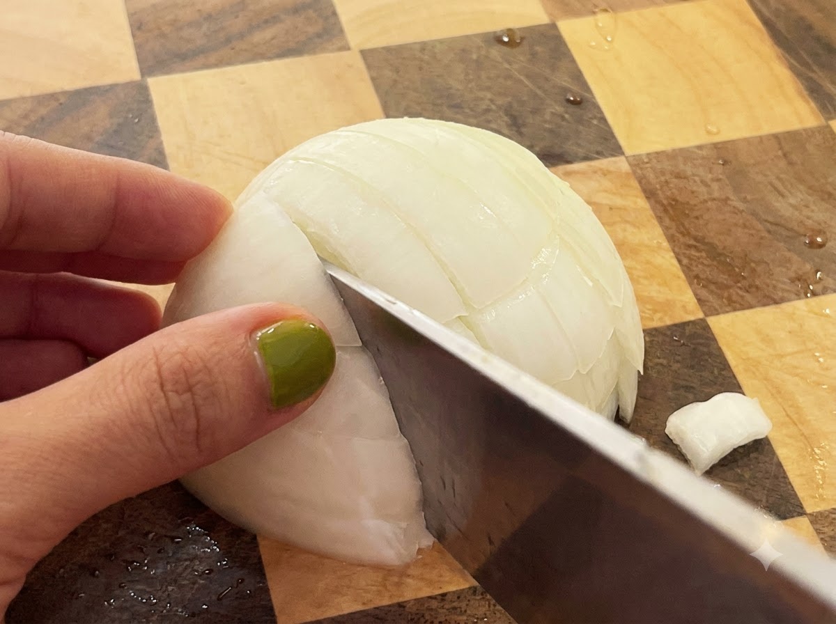 A knife slicing through a halved white onion on a checkered wooden cutting board.