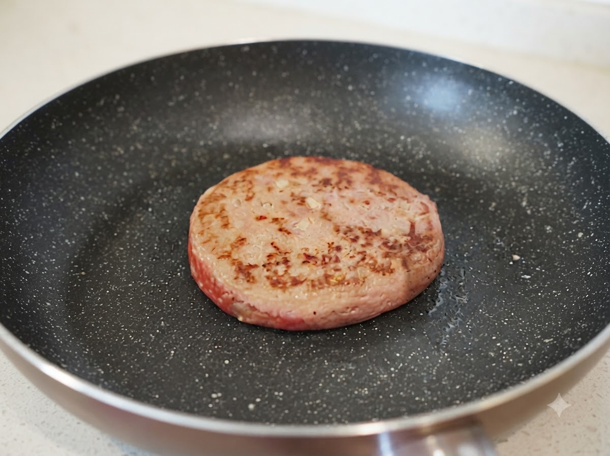 A small stream of water being poured into a frying pan next to a partially browned hamburg patty.