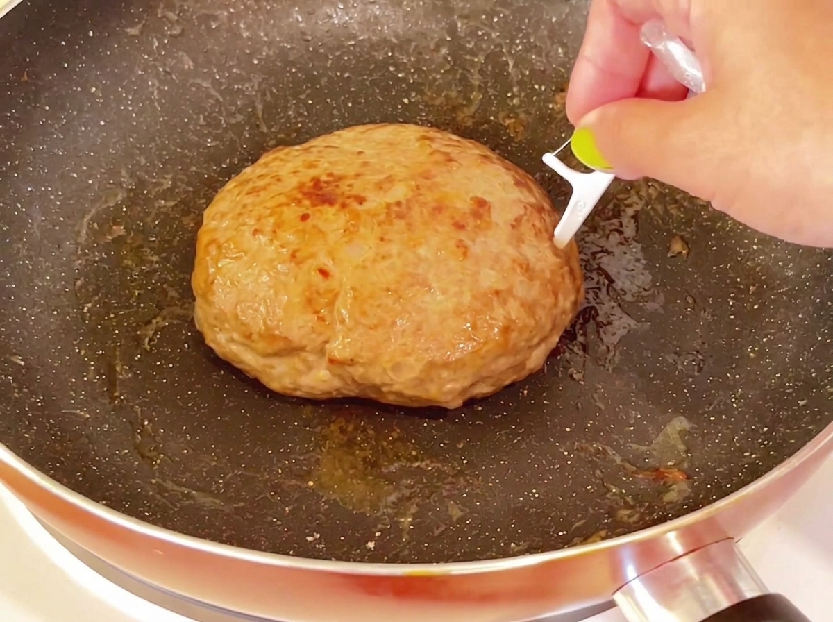 A toothpick being inserted into the top surface of a thick, cooked hamburger patty in a pan.