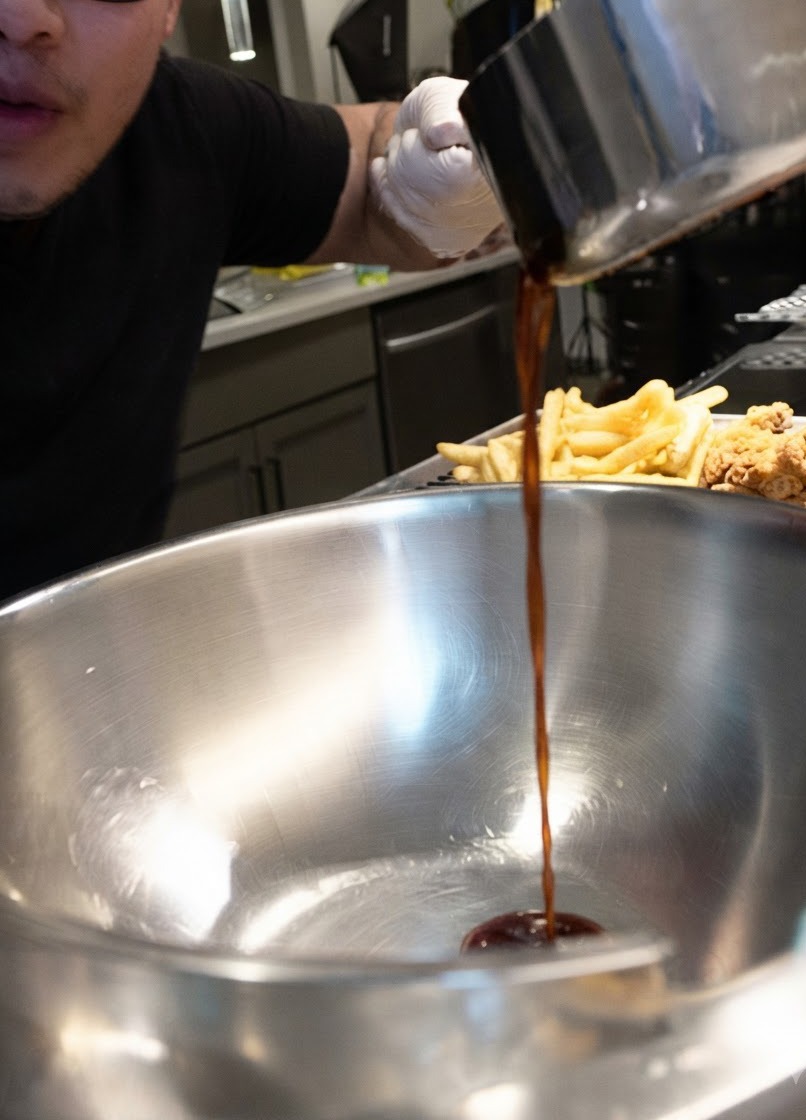 Dark sauce being poured in a steady stream from a saucepan into a large stainless steel bowl next to fried chicken pieces.