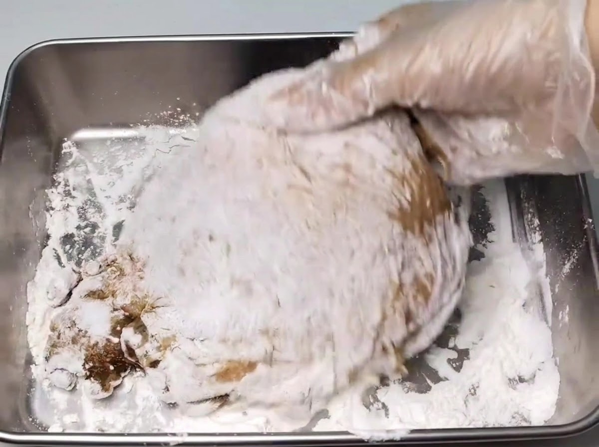 A hand in a glove coating a marinated chicken leg with a layer of white starch inside a metal tray.