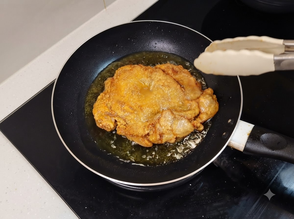 A golden brown fried chicken leg sizzling in an oily frying pan with kitchen tongs nearby.