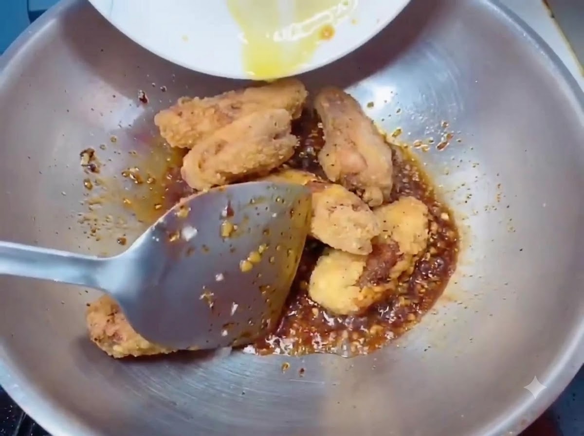 Crispy golden fried chicken wings being transferred into the wok with the sauce using a spatula.