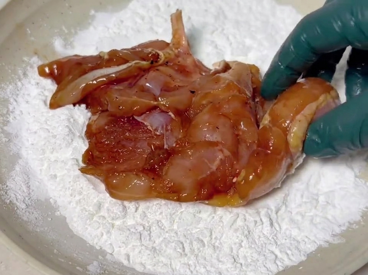Marinated chicken pieces being coated in a white layer of cornstarch in a shallow dish.