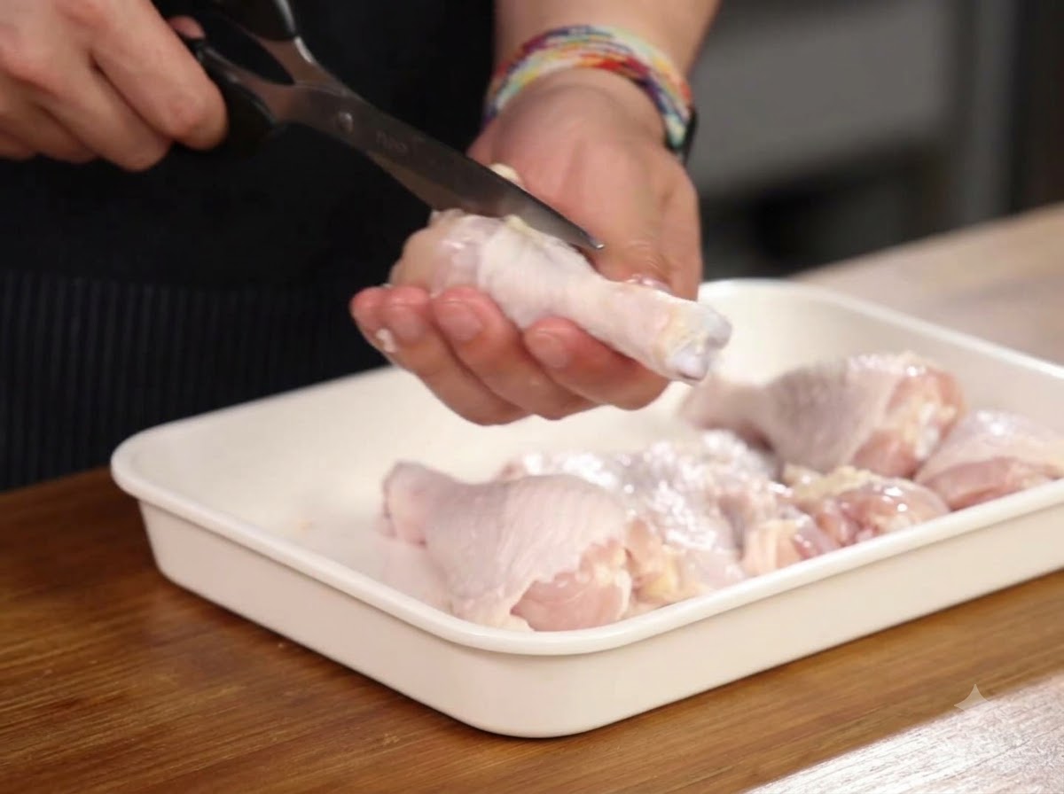 Hands using kitchen scissors to cut around the bone of a chicken drumstick inside a white tray.