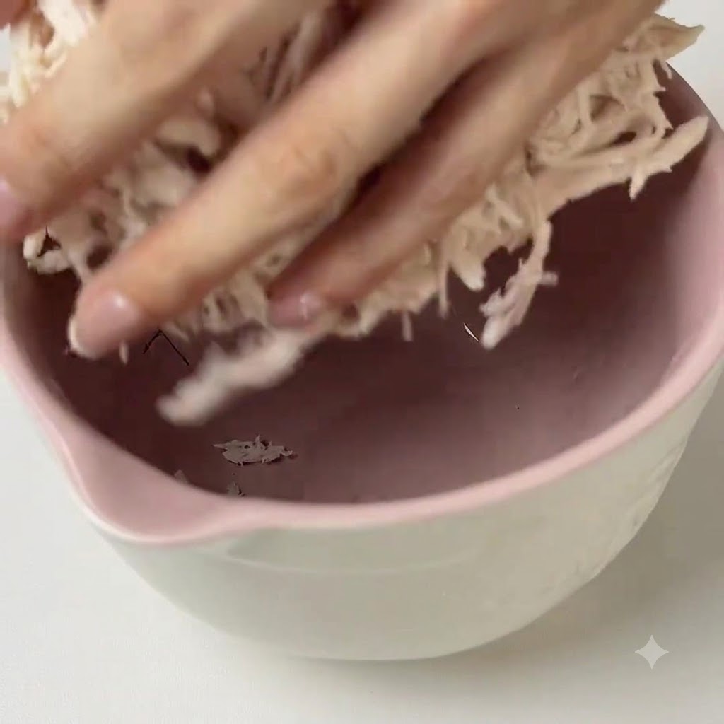 Hands mixing green pesto sauce into shredded chicken inside a pink mixing bowl.