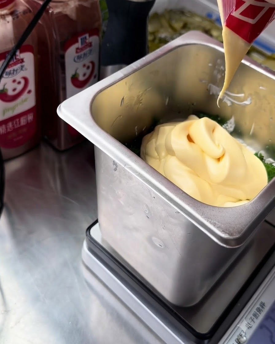 A stream of rich yellow egg yolk mayonnaise being squeezed from a red and white packet into the stainless steel container holding the tartar sauce ingredients.